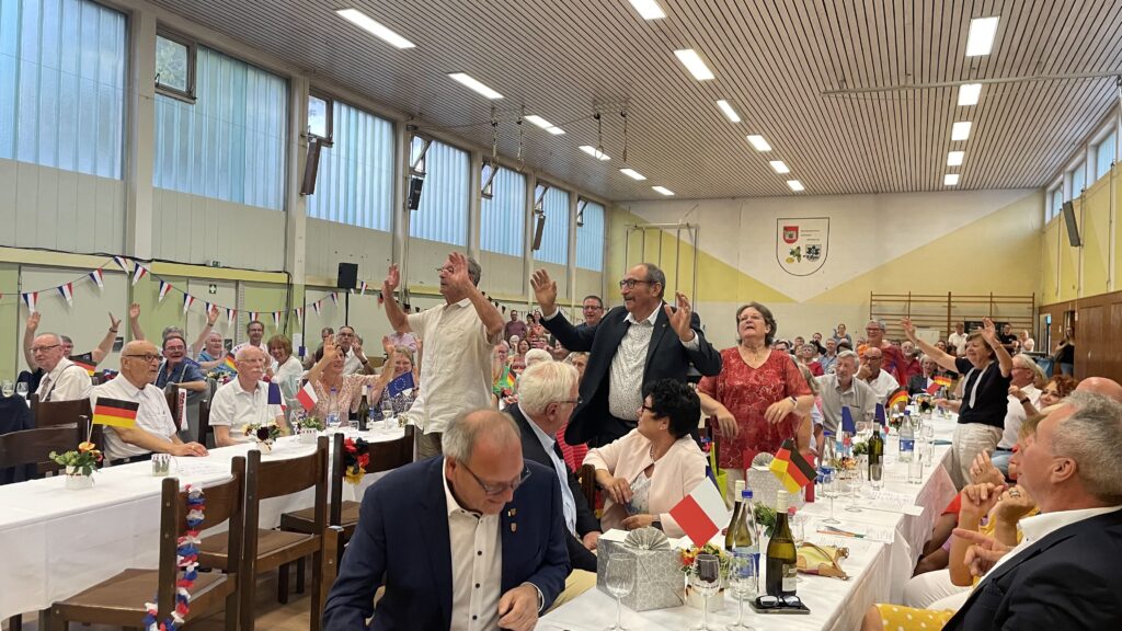 Blick in eine Halle, An langen Tischen sitzen Menschen, einige stehen und heben gerade die Hände.
