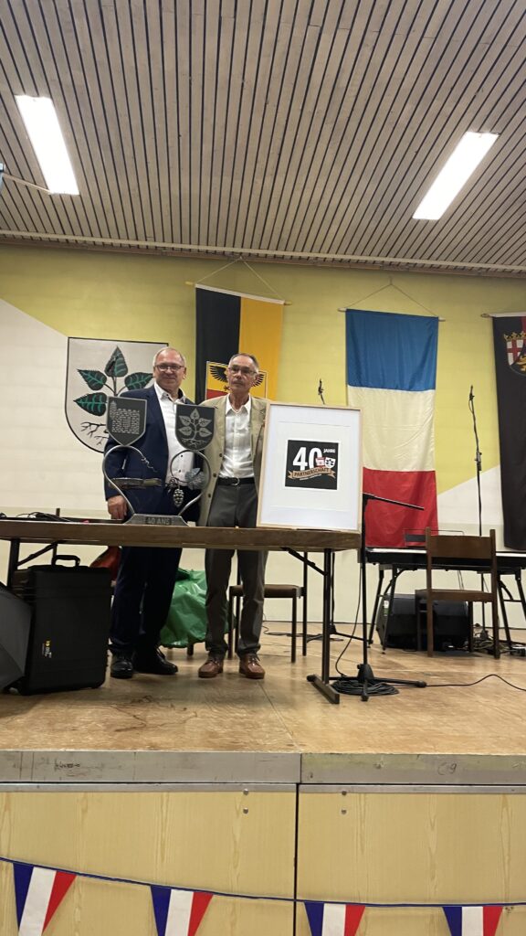 Zwei Männer an einem Tisch auf einer Bühne. Auf dem Tisch eine metallene Skulptur mit zwei Ortswappen sowie einem stilisierten Korkenzieher und einer Traube, darunter der Schriftzug "40 ans". Rechts davon ein gerahmtes Bild mit dem Logo 40 Jahre Partnerschaft.